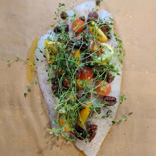 Photo of fish on parchment paper with olives, tomatoes, fresh thyme, capers, photo links to the Private Dinners and Events webpage.